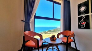 A room with two orange chairs, a small round table with a fruit bowl, and a large window offering a view of the sea and landscape.