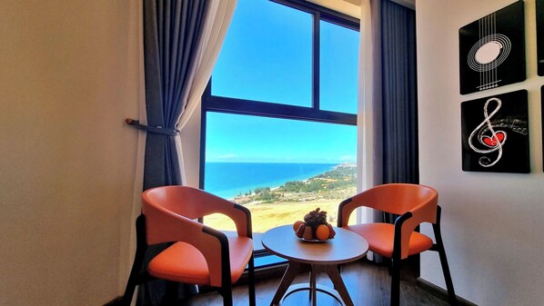 A room with two orange chairs, a small round table with a fruit bowl, and a large window offering a view of the sea and landscape.