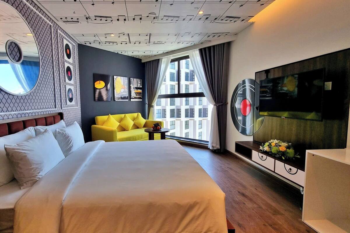 A modern hotel room with a large bed, a yellow sofa, a TV, and a large window with a view of buildings.
