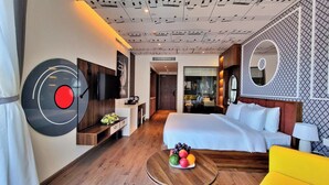 A modern hotel room with a large bed, a flat-screen TV, a record player, and a fruit bowl on a wooden table.
