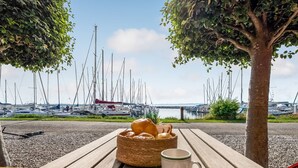 A wooden table with a basket of bread, a mug, and a plate with a knife and spoon. In the background, there are sailboats docked at a marina. - "Ulvi" - 15m from the sea by Interhome (Ebeltoft)