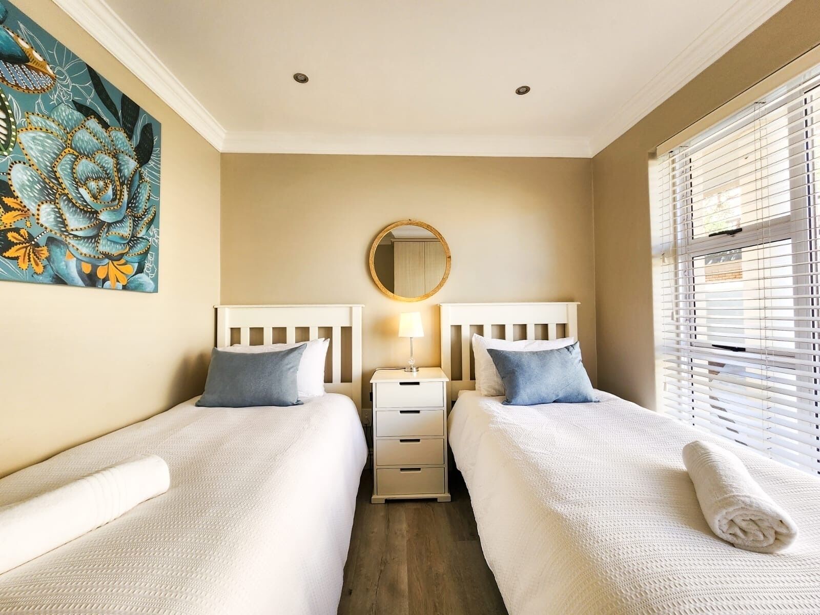 Quarto de hotel com duas camas, um espelho redondo, mesa de cabeceira com abajur e janela com persianas.