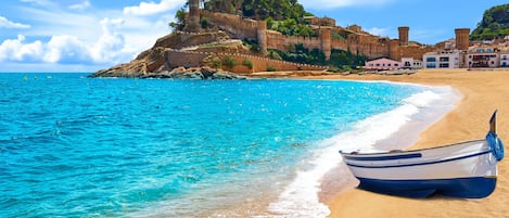A beach with a boat on the sand, a rocky cliff with buildings, and clear blue water.