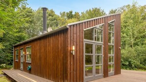 Une cabane en bois dotée de grandes fenêtres, d’une cheminée et d’une terrasse.
