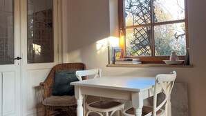 A room with a wooden ceiling, a white table with four chairs, a wicker chair with a cushion, a window with a view of trees, and a door with glass panels.