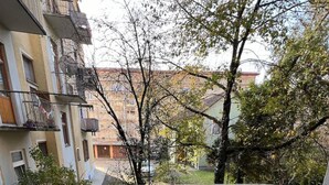 Un balcón con suelo de baldosas, baranda de metal y vista a edificios y árboles vecinos.