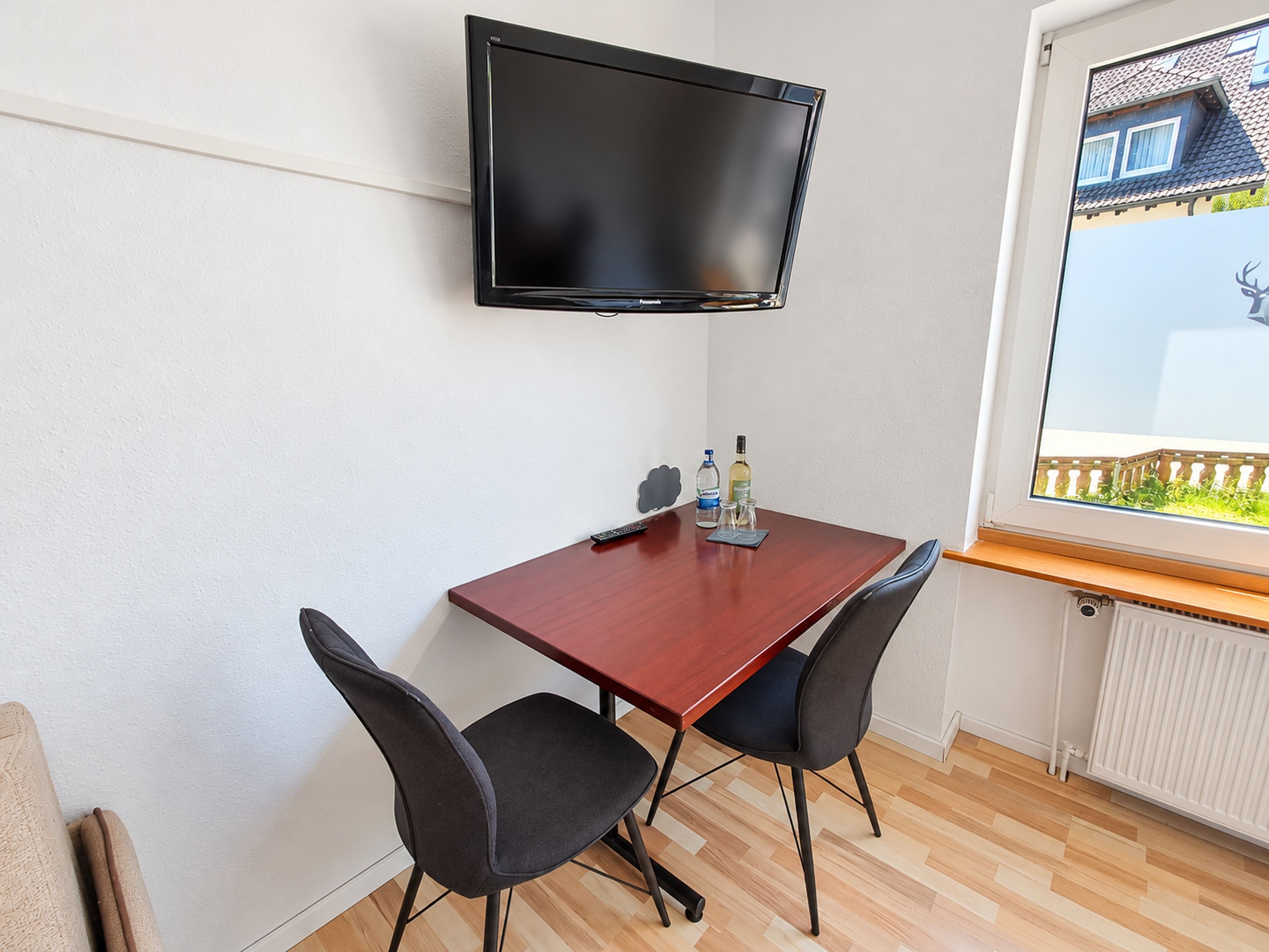 A room with a TV mounted on the wall, a wooden table with two chairs, a window with a view of a house, and a radiator.