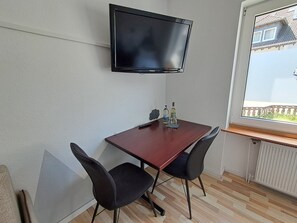 A small room with a TV mounted on the wall, a wooden table with two chairs, and a window with a view of a building outside.