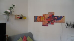 A living room with a grey sofa, green and red cushions, a flat-screen TV, a bookshelf with books and a plant, and a wall with abstract art. - Appartamentino vicino alla Stazione ed al Centro (Padova)