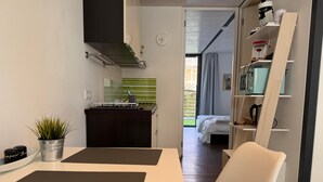 Eine moderne Küche mit Herd, Mikrowelle und Kaffeemaschine. Ein Essbereich mit Tisch und Stühlen ist vorhanden. Im Hintergrund ist ein Schlafzimmer mit Bett und Fenster zu sehen.