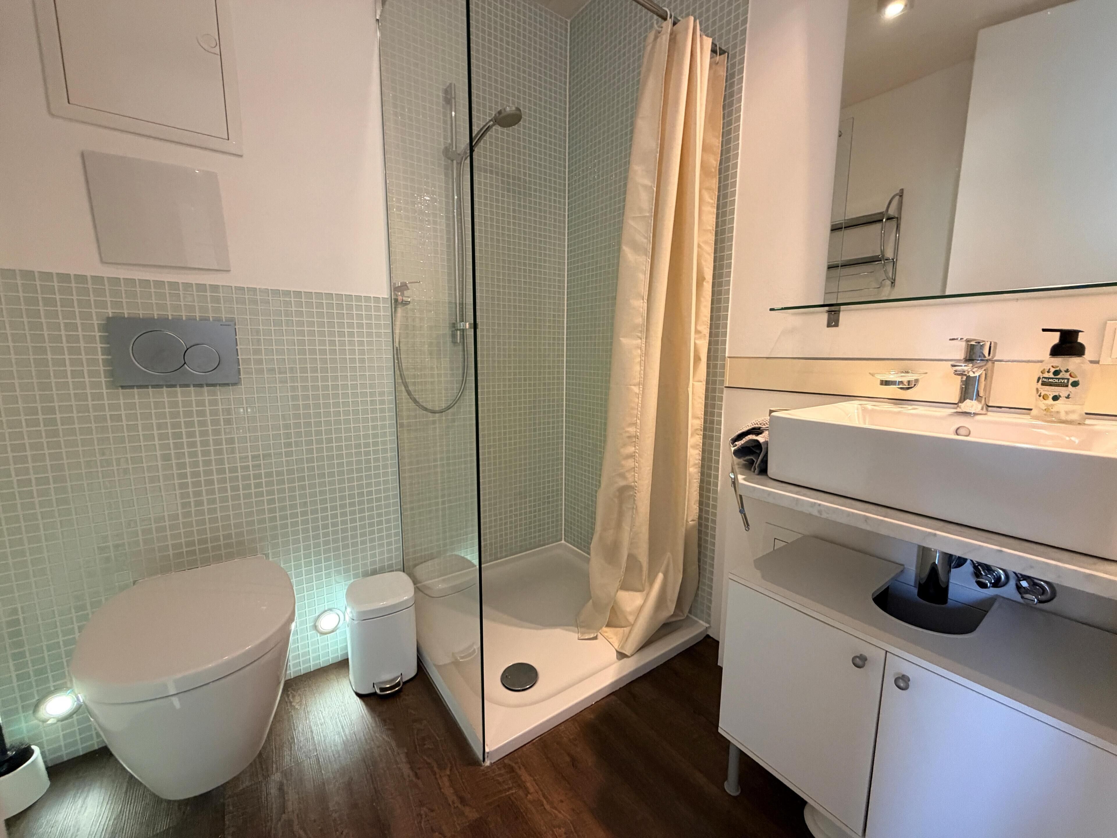 A bathroom with a glass-enclosed shower, a toilet, a sink with a mirror, and a soap dispenser.