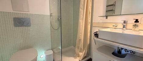 A bathroom with a glass-enclosed shower, a toilet, a sink with a mirror, and a soap dispenser.