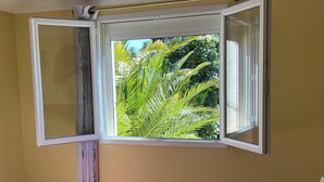 A room with a bed, a window with an open view of palm trees, and a wall socket. - Vacation home with pool (Agde)