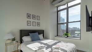 Un dormitorio moderno con una cama grande, una mesita de noche con lámpara, una ventana con vista a la ciudad y obras de arte enmarcadas en la pared.