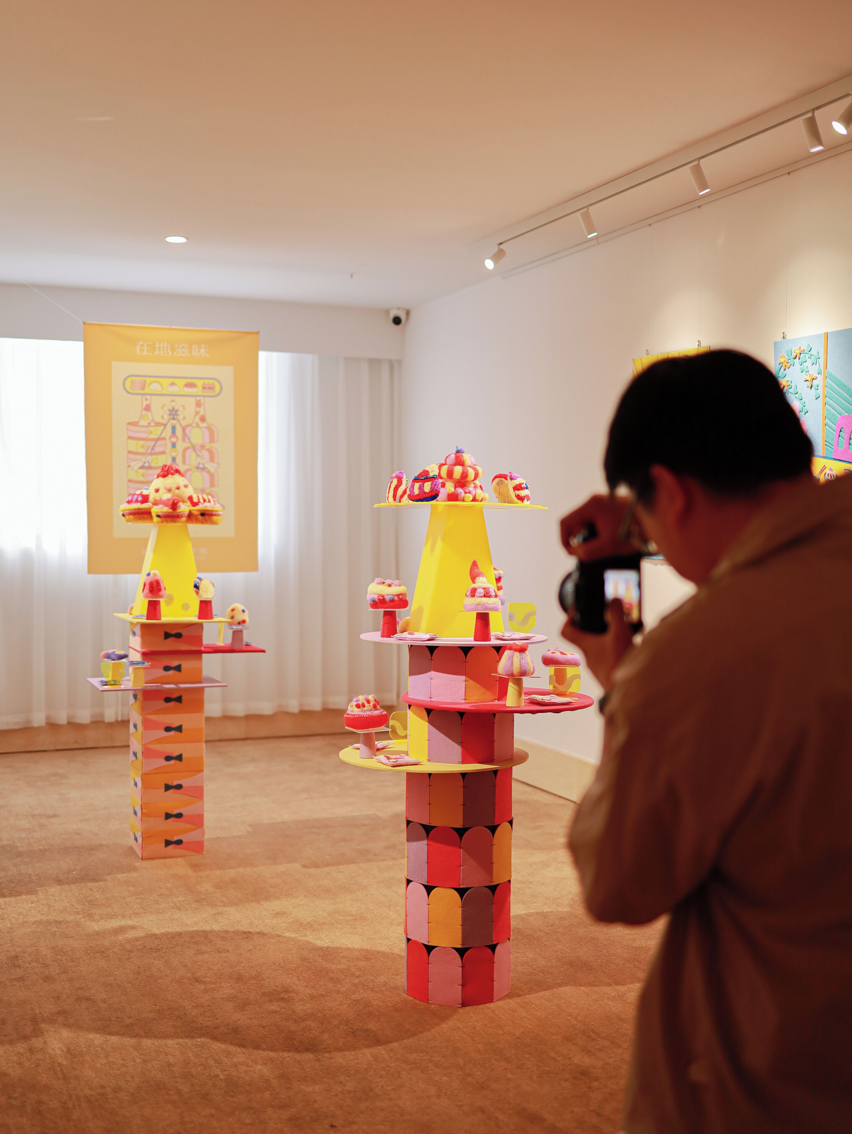 A room with a carpeted floor and a white wall with a poster. There are two yellow and red display stands with cupcakes on them.