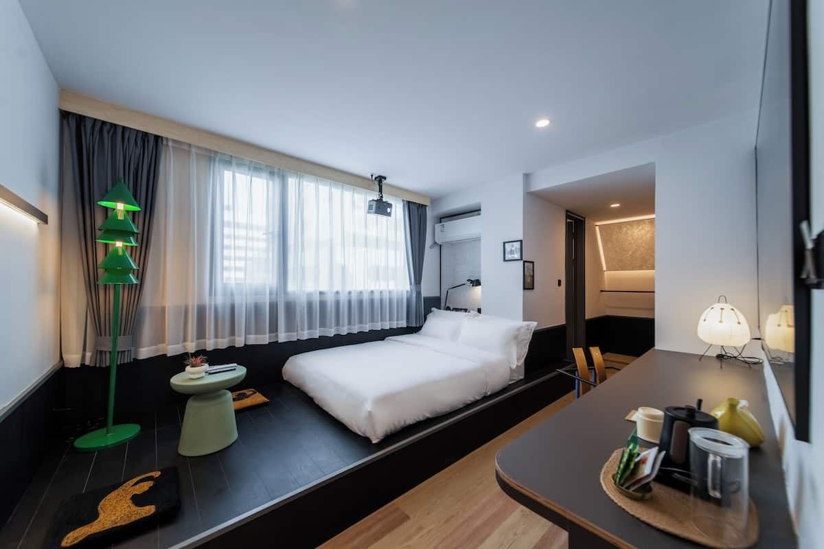 Une chambre d’hôtel moderne, équipée d’un lit, d’une table de chevet, d’une lampe et offrant une vue sur la ville grâce à de grandes fenêtres.