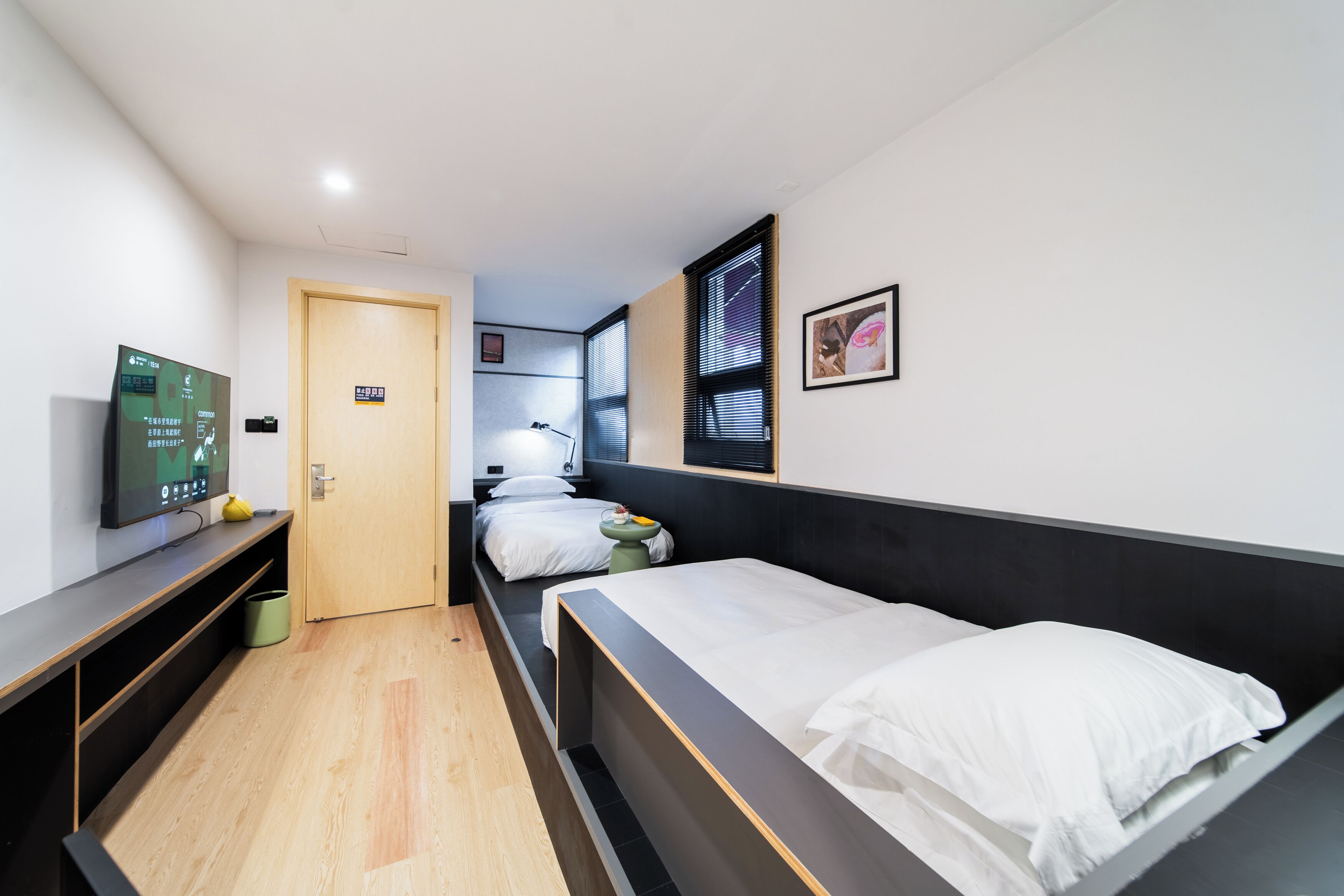 A modern hotel room with a wooden floor, a bed with white bedding, a desk with a flat-screen TV, a small bedside table, and a window with blinds.