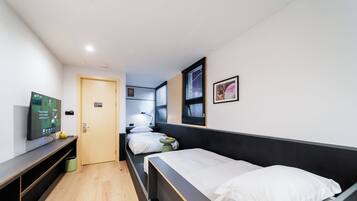 A modern hotel room with a wooden floor, a bed with white bedding, a desk with a flat-screen TV, a small bedside table, and a window with blinds.
