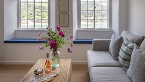 Une pièce avec un canapé, un siège de fenêtre, un vase rempli de fleurs et une bouteille posée sur une table.