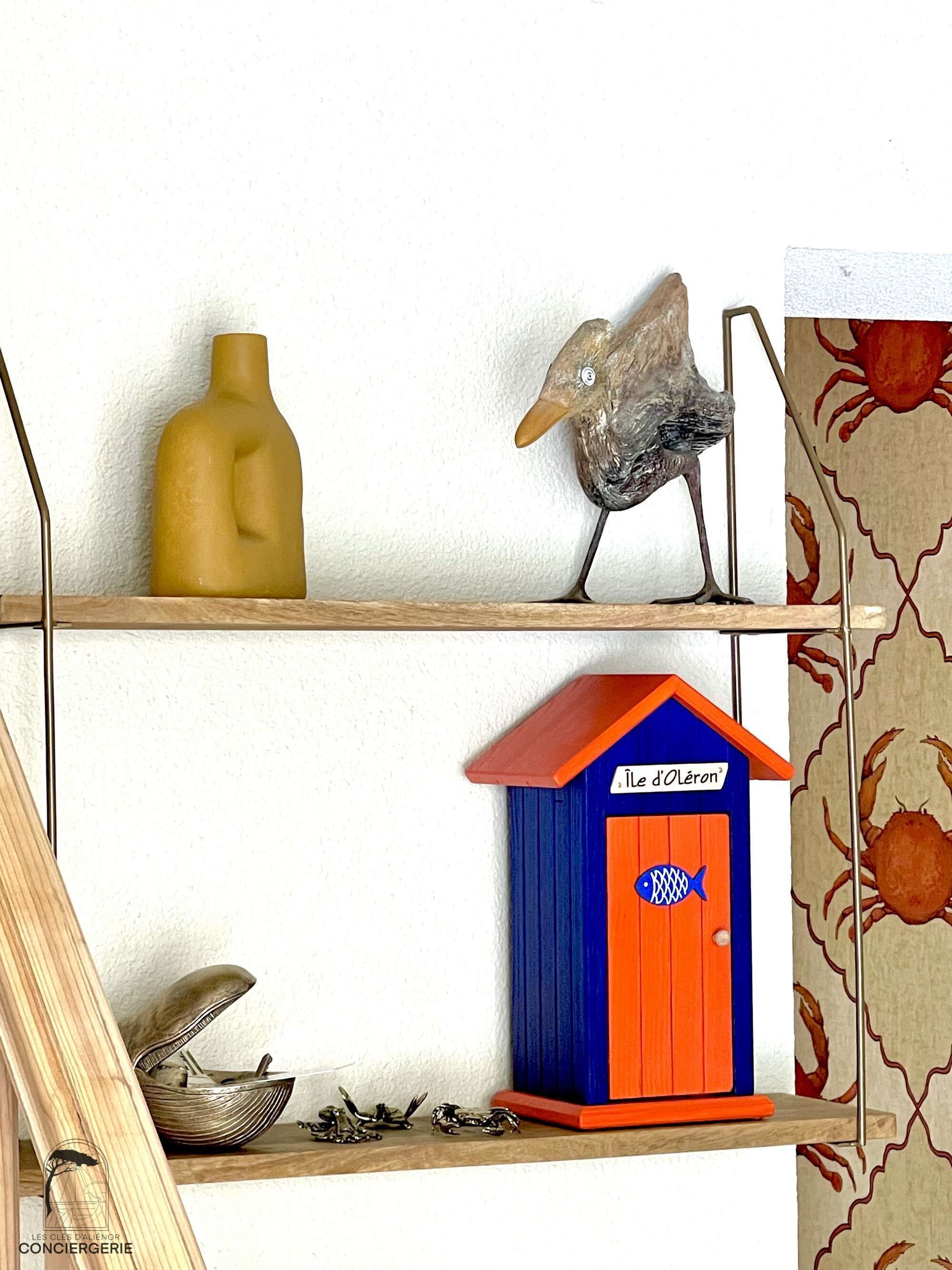 Een plank met een gele vaas, een beeld van een vogel, een blauw-oranje vis huisje en een schaal met krabben.