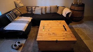 A cozy living room with a wooden coffee table, a black sofa with multiple cushions, a wooden barrel, and a wall-mounted light fixture. - Charming comfort room (Fénols)