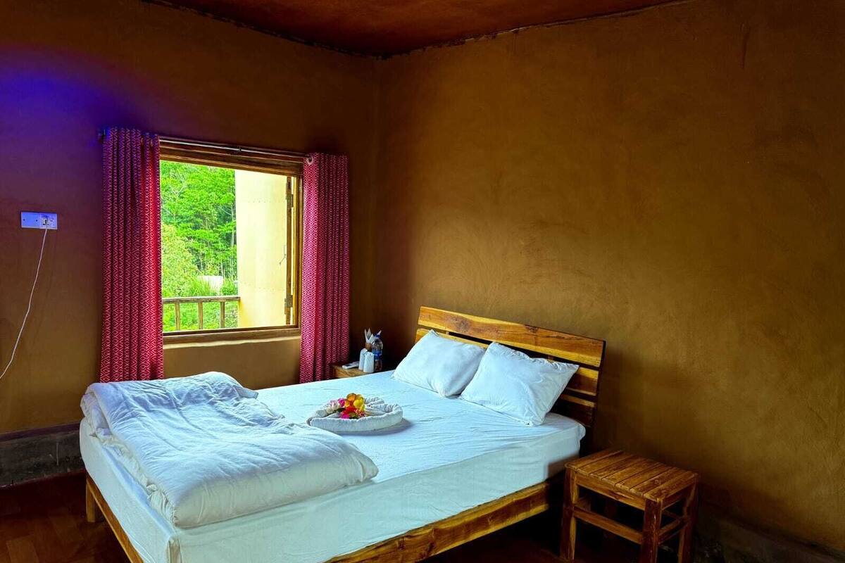 A wooden bed with white linens and pillows, a small wooden stool, a window with red curtains, and a view of greenery outside.