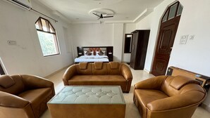A spacious living room with a brown leather sofa set, a glass coffee table, and a bed in the background. - Velora Inn  (Varanasi)