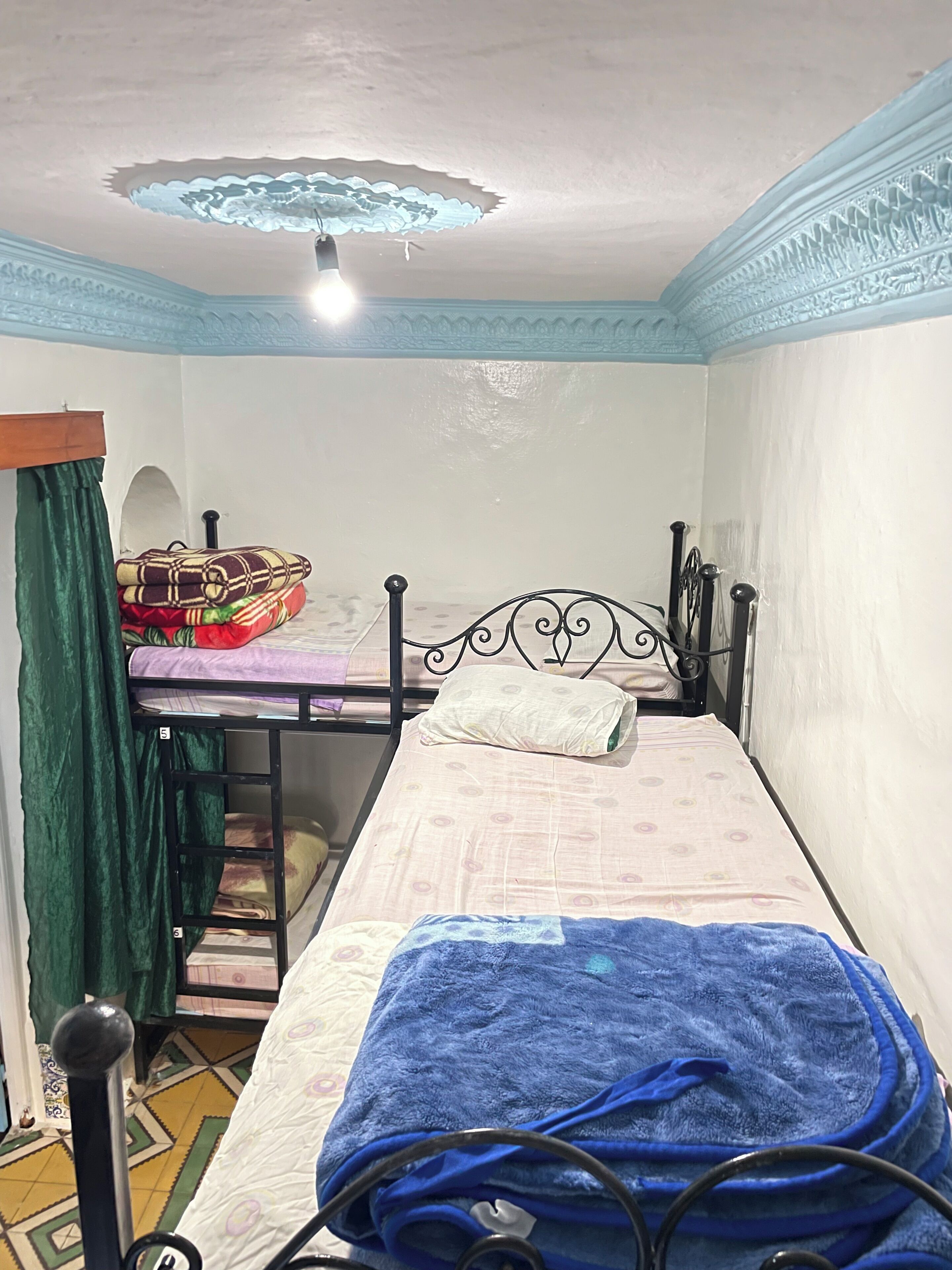 Una cama individual con una colcha azul, una cortina verde y un aplique decorativo en el techo.