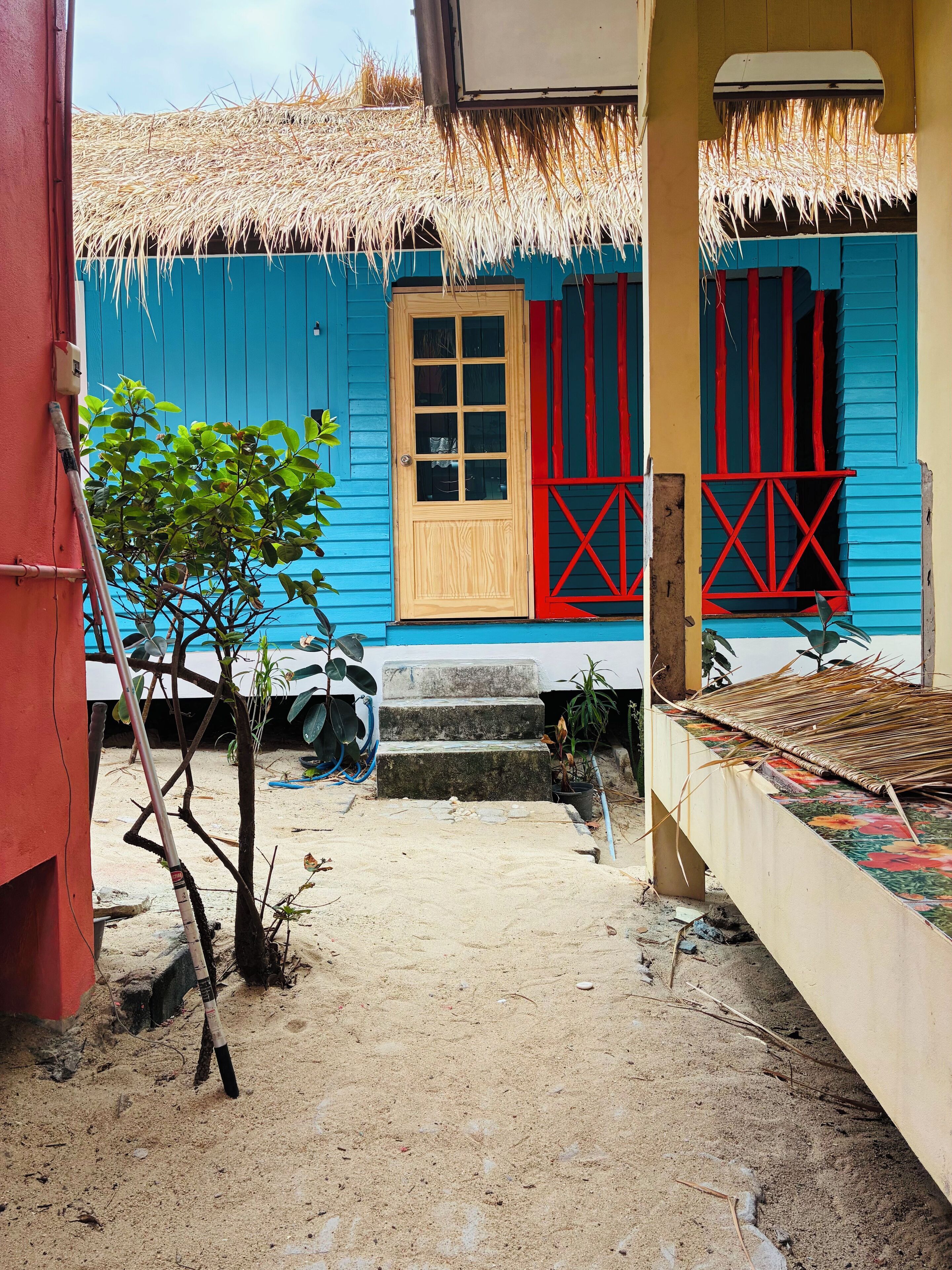 A thatched-roof building with a wooden door and red window shutters, a small staircase leading to the entrance, and a sandy ground area with a plant and a broom.