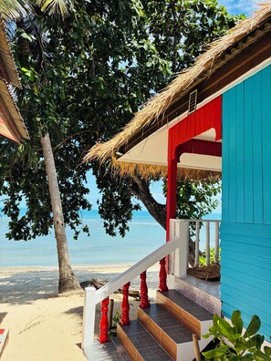 Un escalier menant à la plage, avec des balustrades rouges, aboutit à un bâtiment bleu doté d’un toit de chaume.