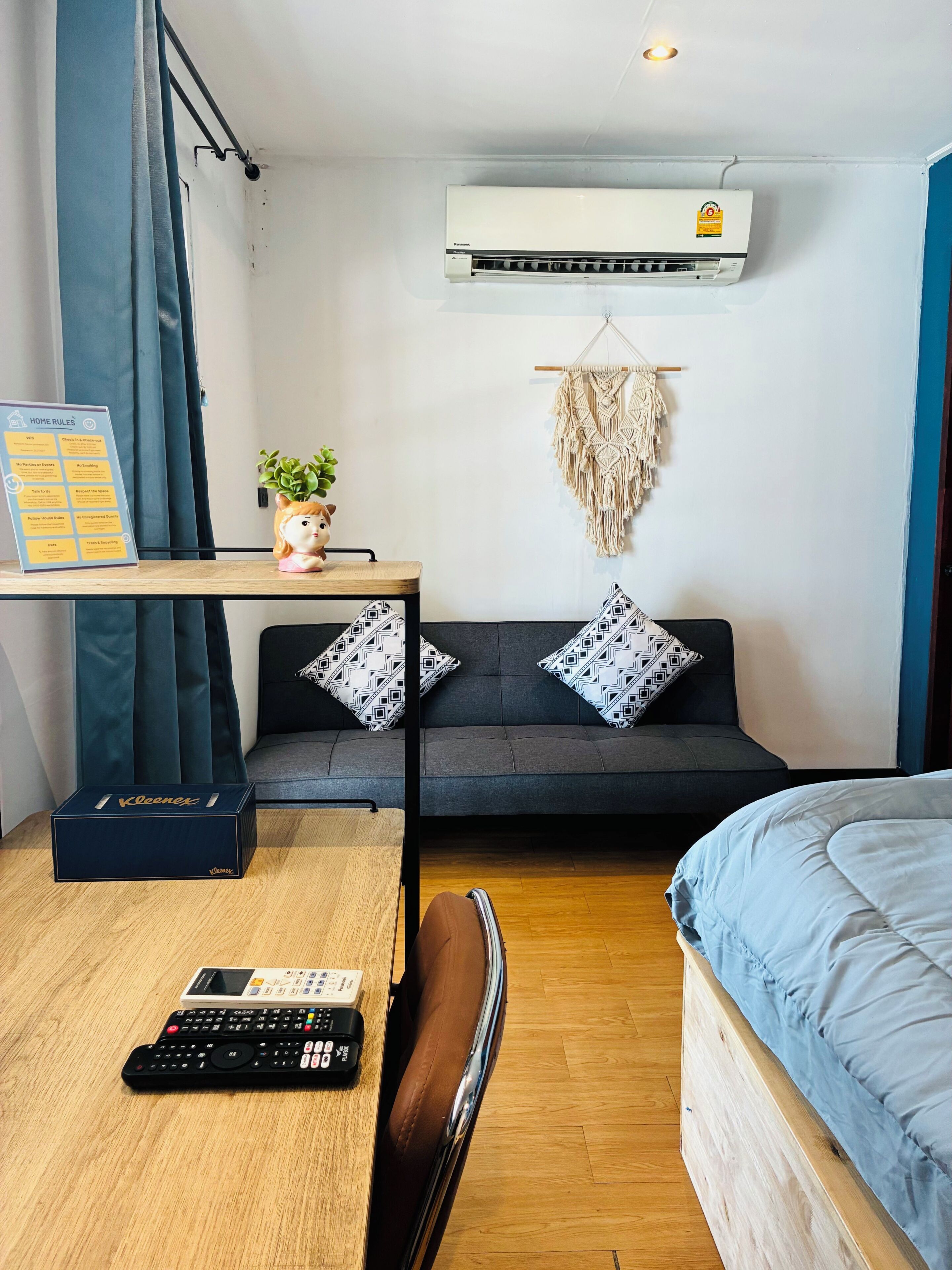 A modern room with a grey sofa, patterned pillows, a macrame wall hanging, a small plant, a desk with a sign, a blue bed, and a wooden floor.
