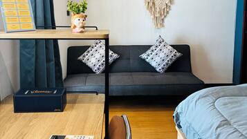 A modern room with a grey sofa, patterned pillows, a macrame wall hanging, a small plant, a desk with a sign, a blue bed, and a wooden floor.