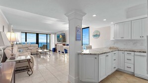 A spacious living area with a large window overlooking the ocean, a kitchen with white cabinetry, and a dining area with a table and chairs.