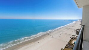 A beach view from a balcony with a clear sky, a sandy beach, and a pier in the distance.