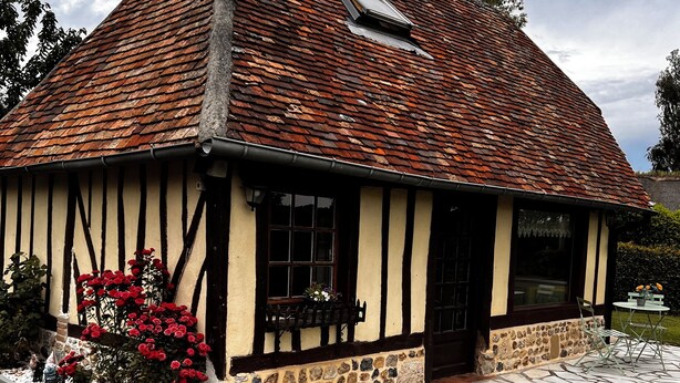 Une maison traditionnelle à colombages, avec des fondations en pierre, un toit à deux versants et un petit jardin agrémenté de fleurs rouges.