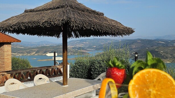 A table with a drink, a straw, and a slice of orange, set against a scenic backdrop with a thatched umbrella and a view of distant hills.
