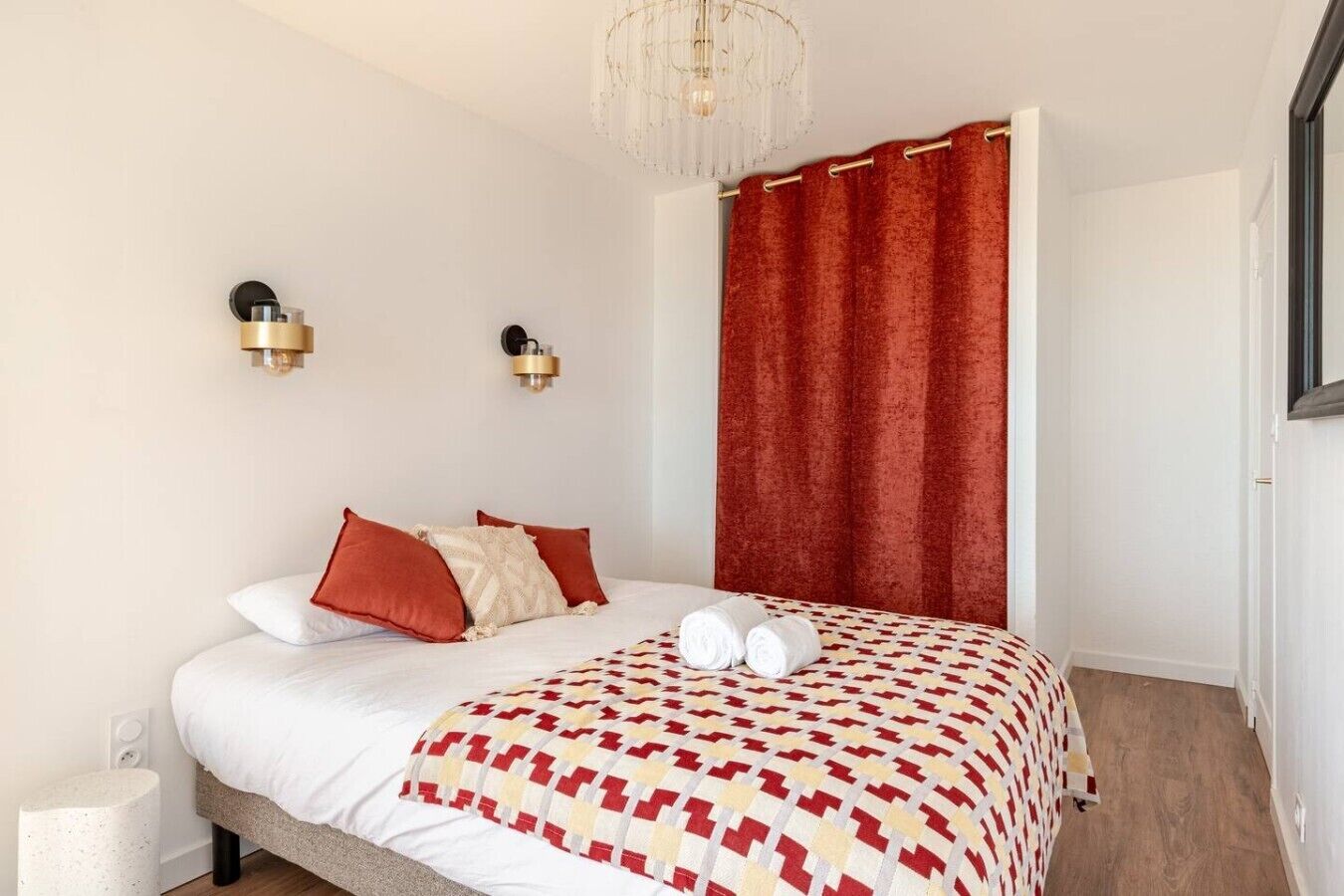 A bedroom with a bed, red curtains, two wall-mounted light fixtures, and a chandelier.