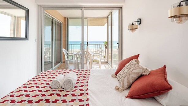 Ein Schlafzimmer mit einem Bett, zwei Kissen, einer Decke, einer Chandelüster, einem Nachttisch und Blick auf das Meer durch eine Schiebetür.