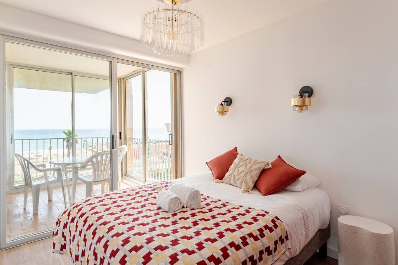 A bedroom with a bed, a chandelier, a balcony with chairs, and a view of the sea.