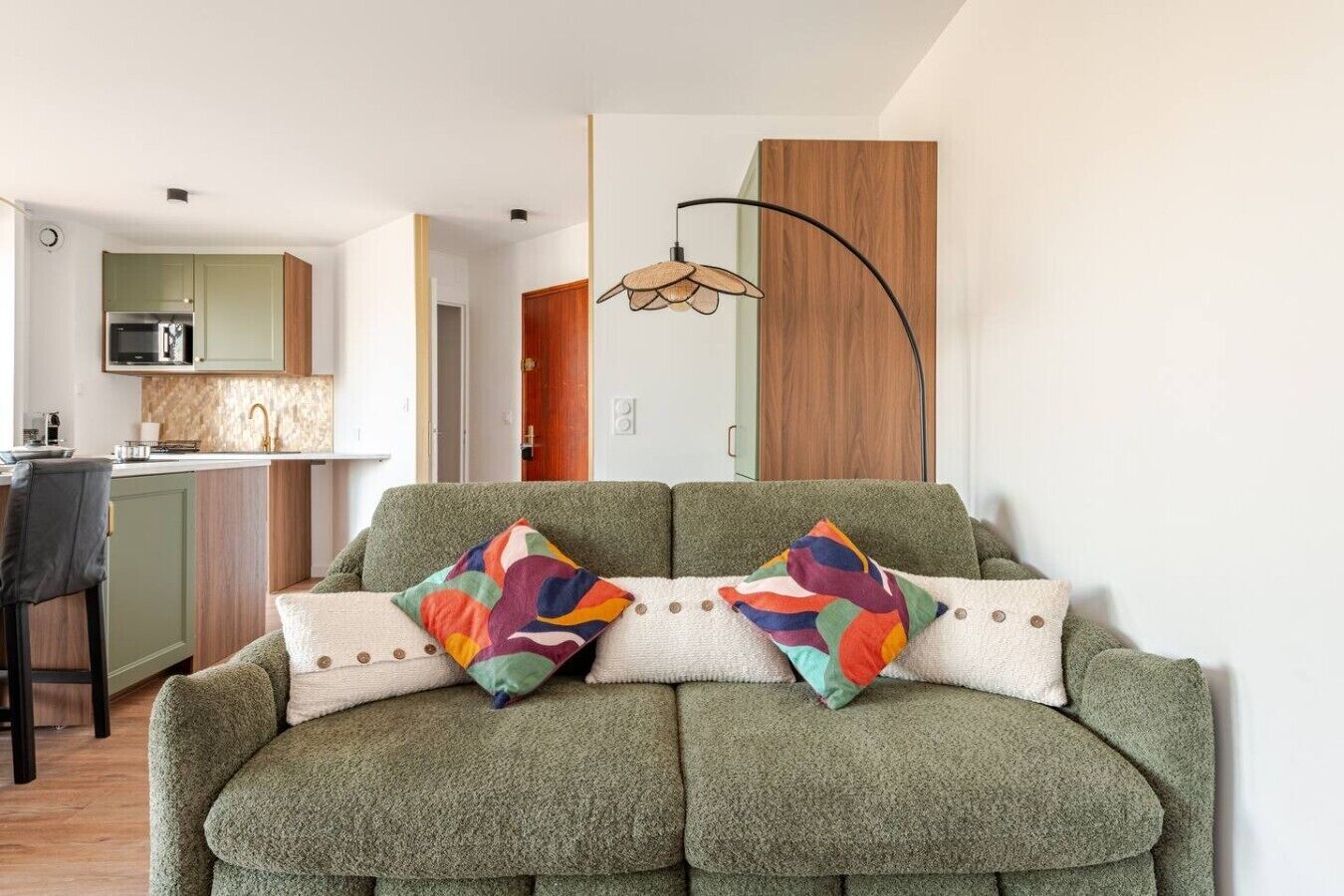 A green sofa with colorful pillows, a kitchen area with green cabinets, and a wooden door.