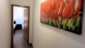 Un couloir orné d’un tableau représentant des tulipes rouges, une porte en bois et, au fond, on aperçoit une pièce avec un lit et une chaise.