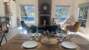 A dining area with a wooden table set for four, a fireplace, and large windows with a view of greenery. - Modern, comfort and serenity in Fitzroy Falls (Fitzroy Falls)