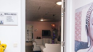 A room with a white couch, a small table, and a wall-mounted shelf. There is a sign on the wall listing amenities such as a lounge, fitness area, massage, and cosmetics.
