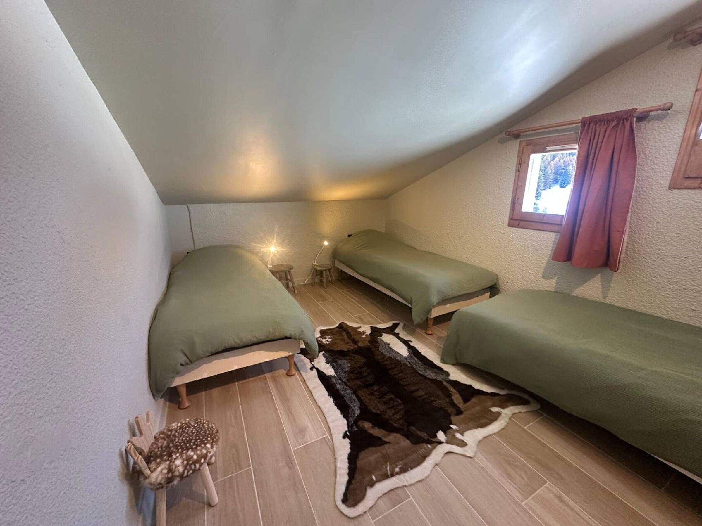 Une petite chambre mansardée, chaleureuse et accueillante, avec deux lits simples, une fenêtre avec des rideaux, un tabouret en bois et un tapis en fourrure sur le sol.