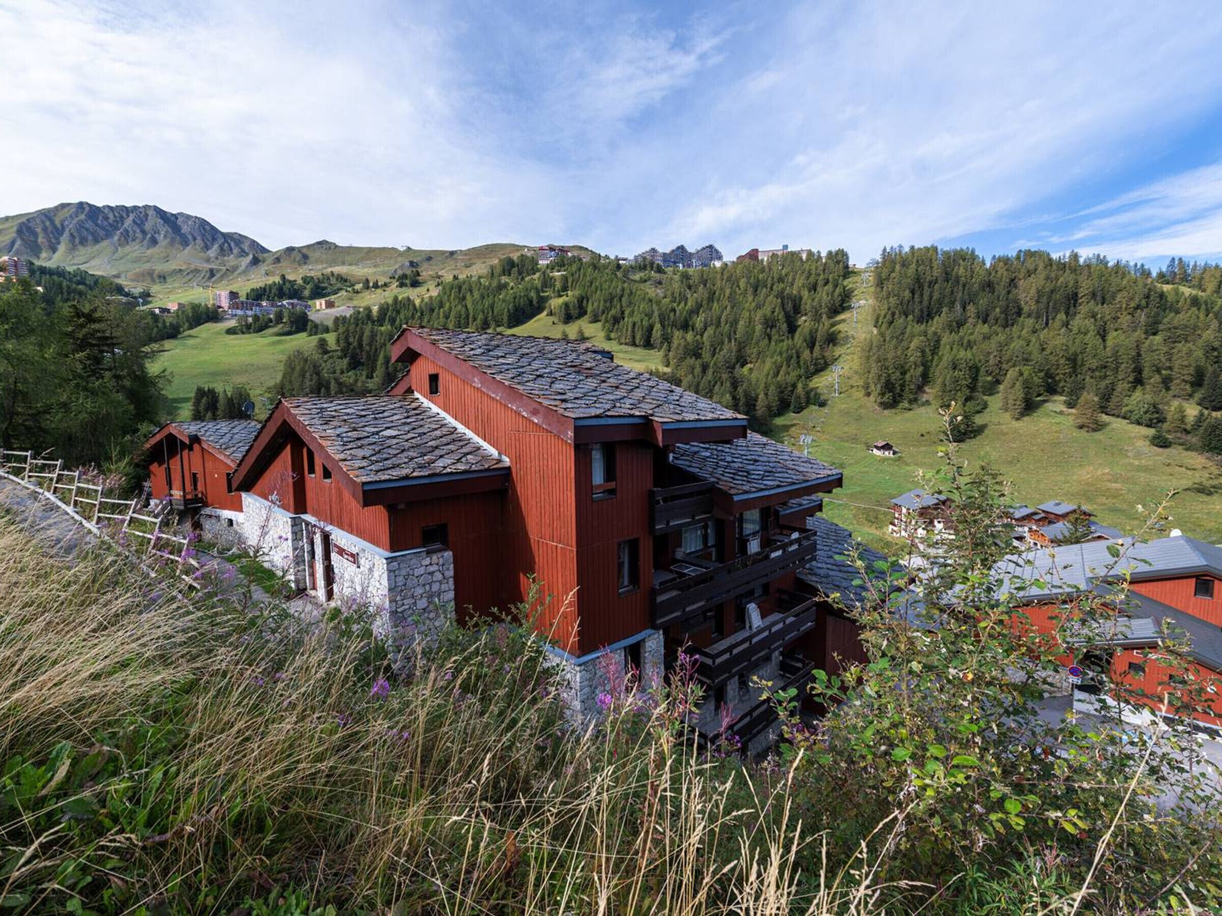 A red wooden building with a stone base, surrounded by greenery and mountains in the background.