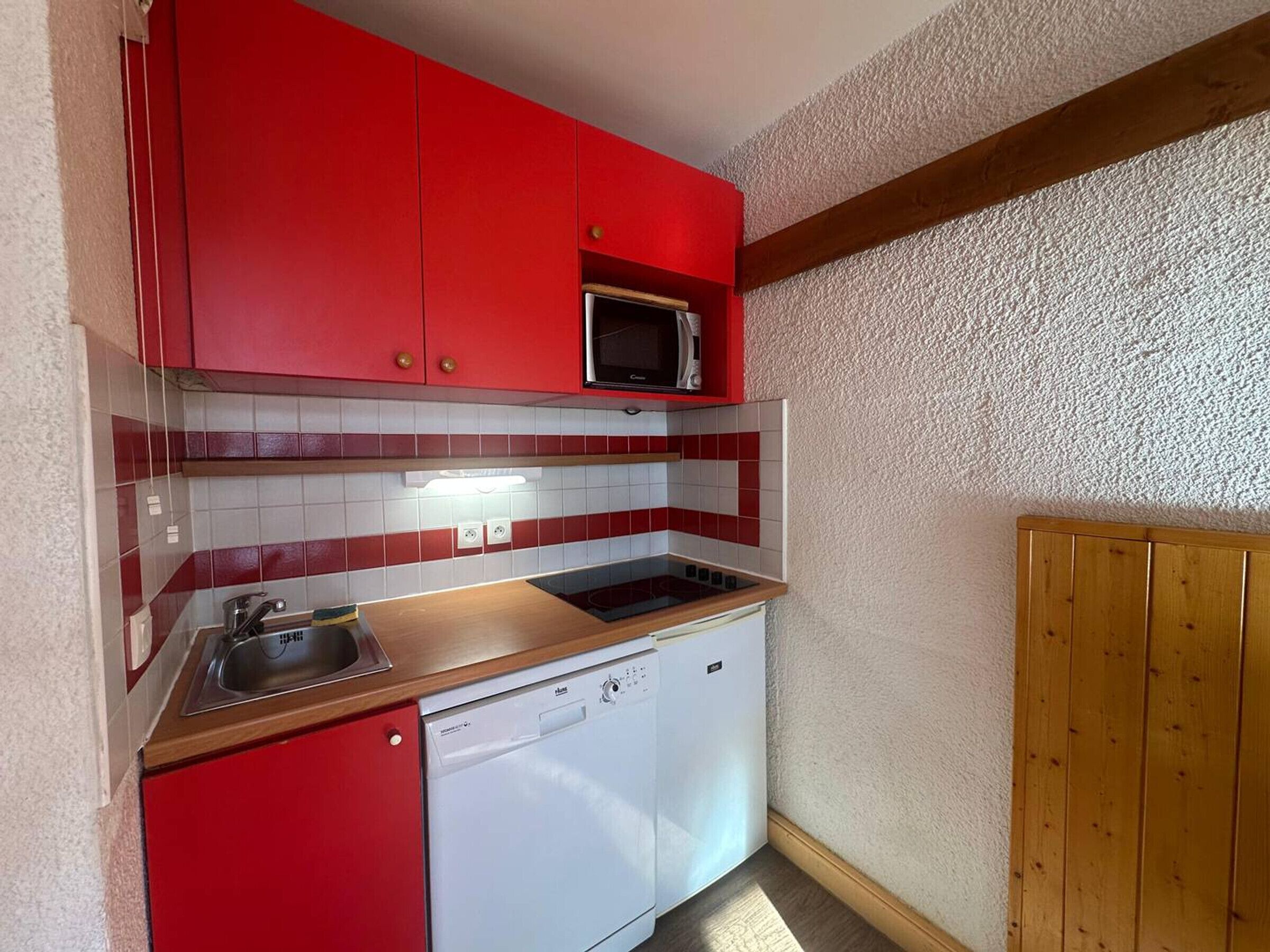 Une cuisine compacte avec des meubles de rangement rouges, un four à micro-ondes, un lave-vaisselle et un mur recouvert de panneaux en bois.