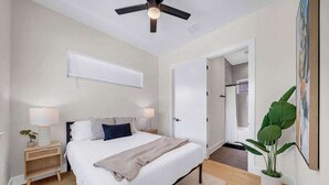 A bedroom with a bed, bedside tables, a ceiling fan, a window with blinds, a door, and a potted plant.