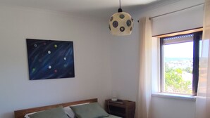 A bedroom with a bed, a nightstand, a lamp, a painting, and a window with a view.