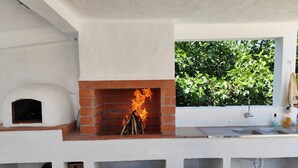 An outdoor kitchen with a brick fireplace, a pizza oven, and a window overlooking greenery.