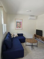 Una sala moderna con un sofá seccional azul, una mesa de centro de madera, un televisor de pantalla plana sobre un mueble de madera y una obra enmarcada en la pared.